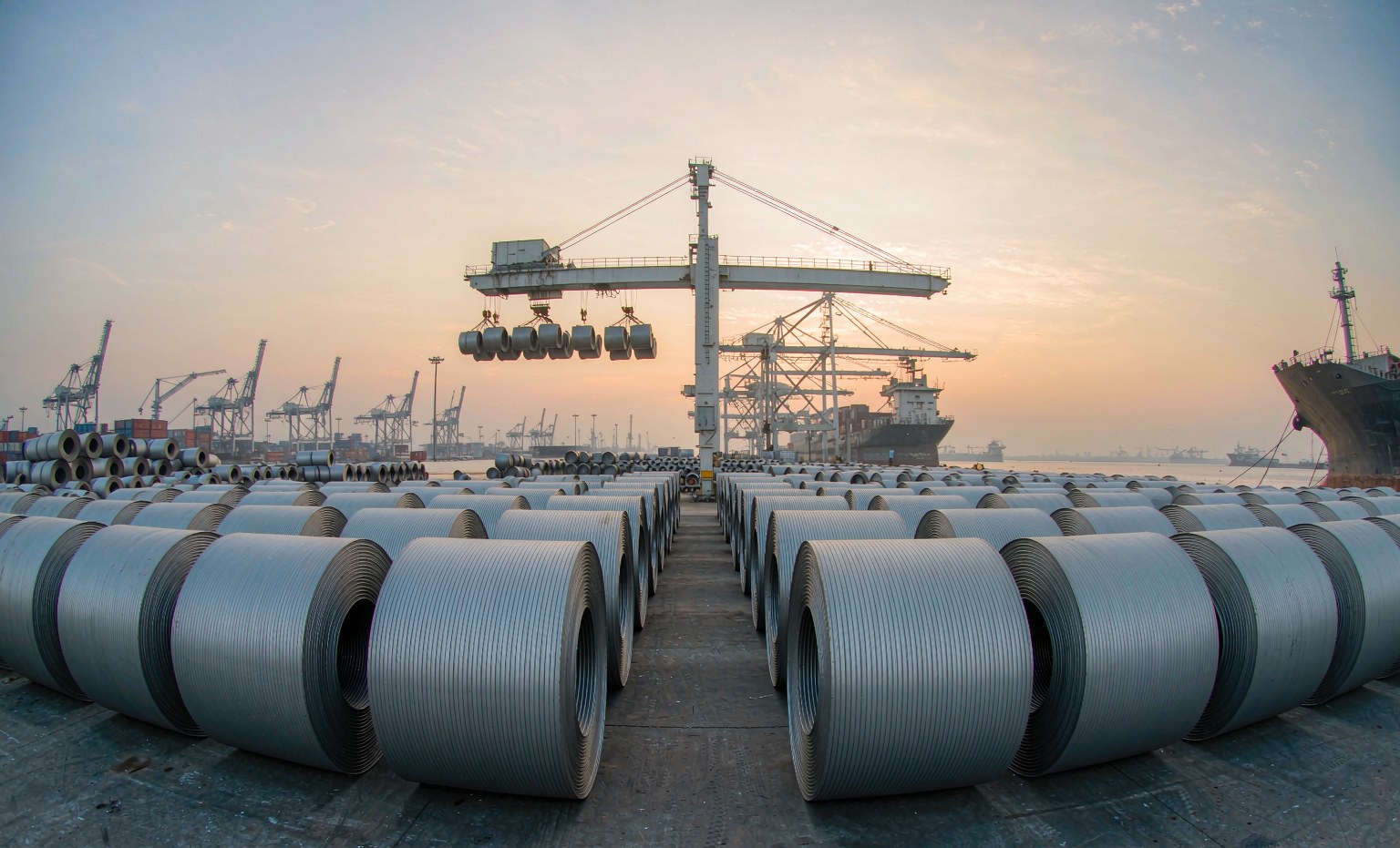 Steel coils stacked at a seaport export terminal with gantry cranes loading a cargo ship at dusk
