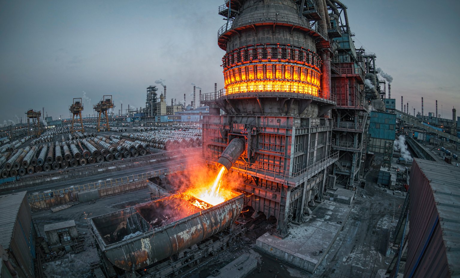 A modern integrated steel plant at dusk with a glowing blast furnace, torpedo ladle car and steel coil yard