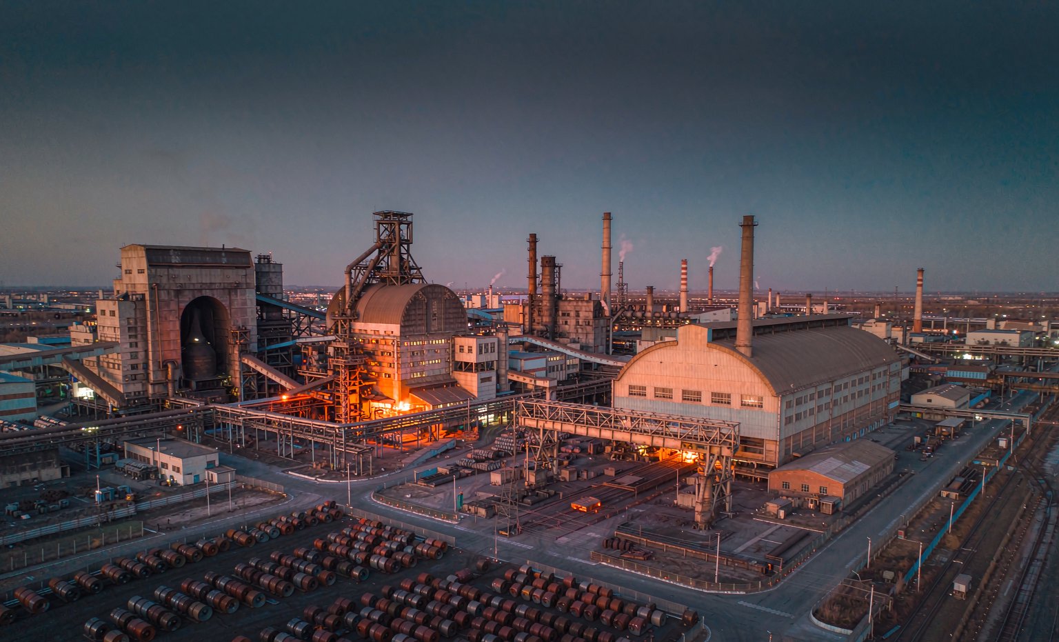 Aerial view of a modern integrated steel plant at dusk with glowing molten steel and stacked coils